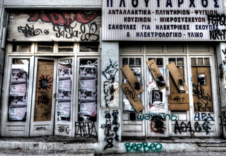 Abandoned graffiti-covered storefront featuring Greek signs and closed doors.