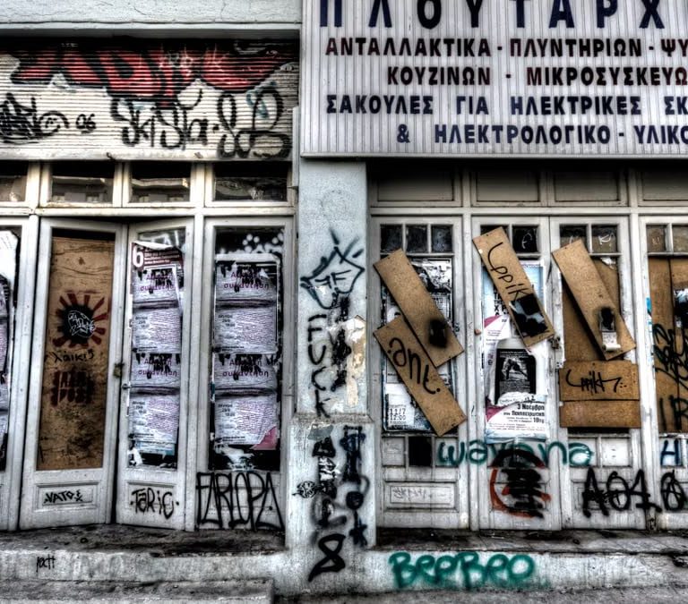 Abandoned graffiti-covered storefront featuring Greek signs and closed doors.